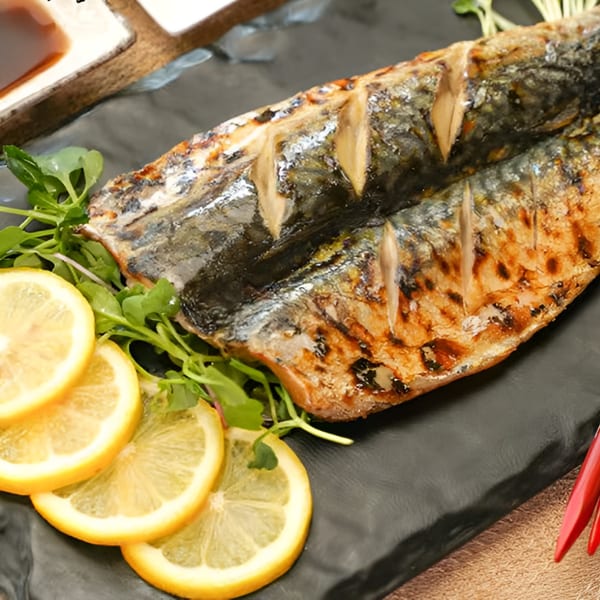 Grilled mackerel with scored skin, garnished with fresh greens and lemon slices on a black plate.
